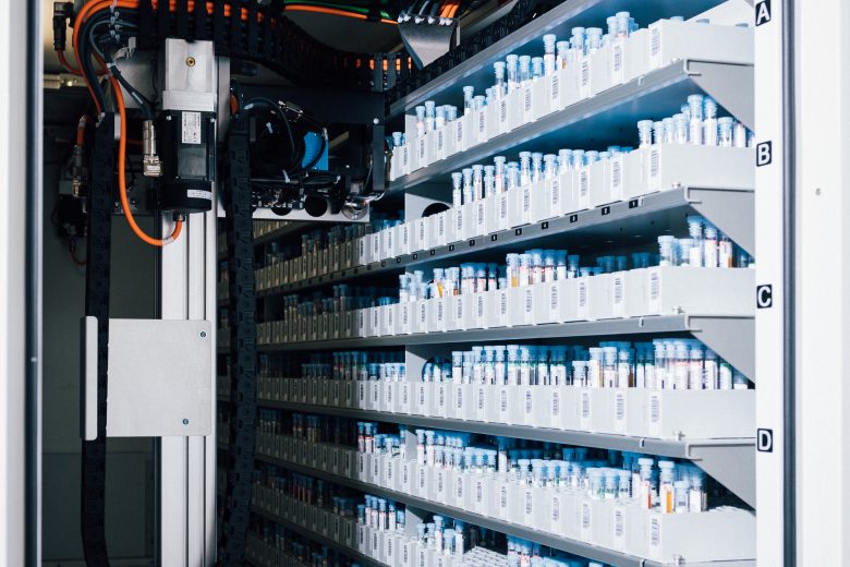 Gerät neben Regal mit zahlreichen transparenten Probenröhrchen mit blauen Deckeln und Barcode-Aufklebern.