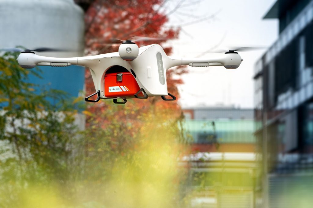 Startende weiße Drohne mit roter Probentransportbox vor dem Zentrallabor von Labor Berlin und Bäumen im Hintergrund.